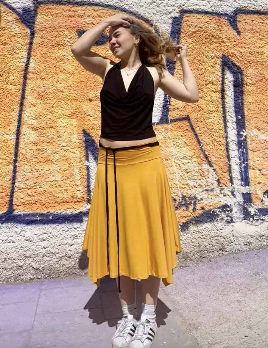 Japamala customer with black Eva top and mustard maxi skirt. Wearing her Japamala outfit in front of a mural of the streets of Gracia, outside the store. 