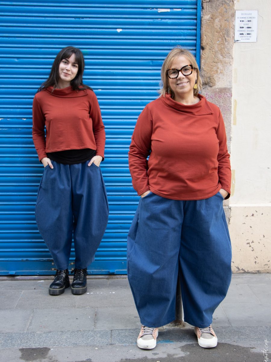 Dos mujeres posan frente a una persiana metálica azul. Ambas llevan un jersey color teja y pantalones anchos denim. La mujer de la derecha usa gafas y zapatillas claras, mientras que la mujer de la izquierda lleva botas negras.