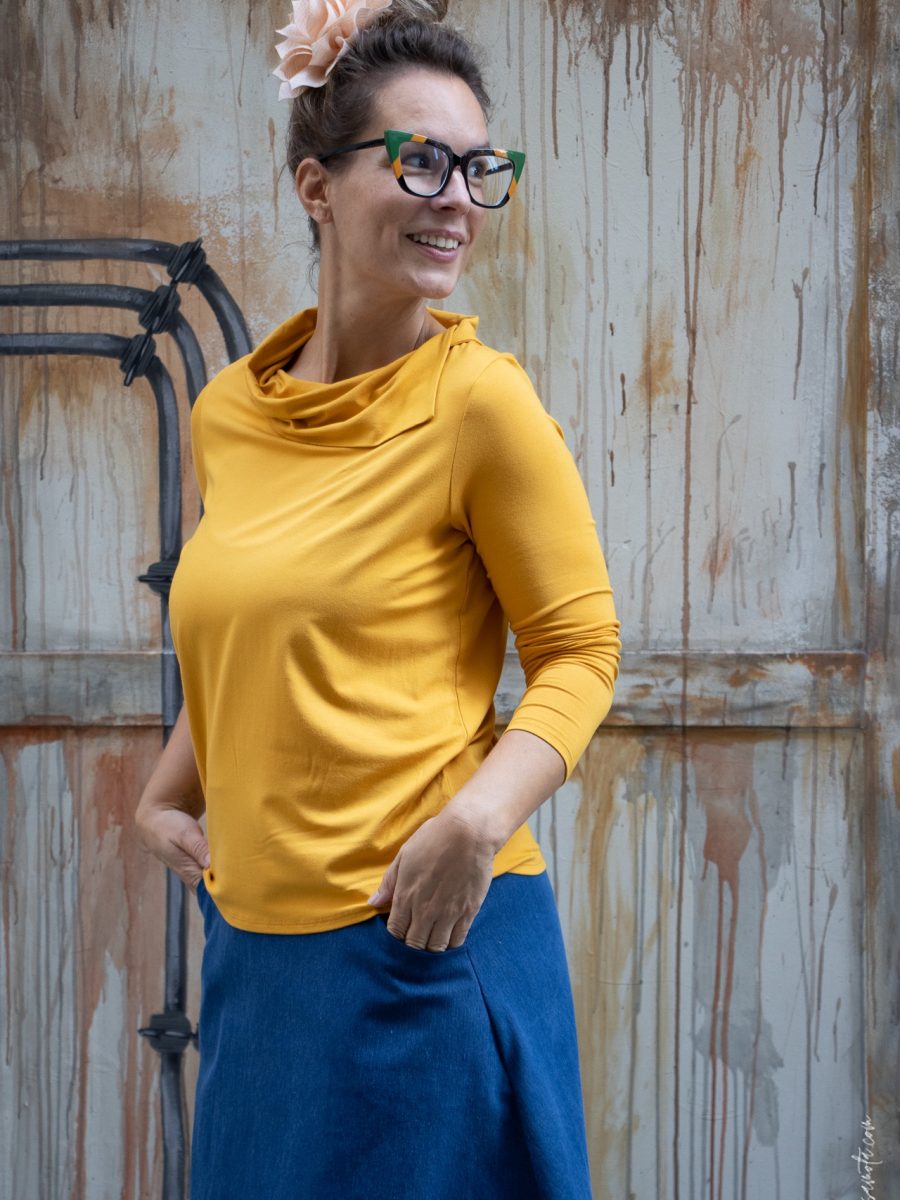 Mujer sonriendo con top leda amarillo mostaza de manga tres cuartos y cuello drapeado, combinando con falda azul. Moda casual y elegante de la colección Leda.