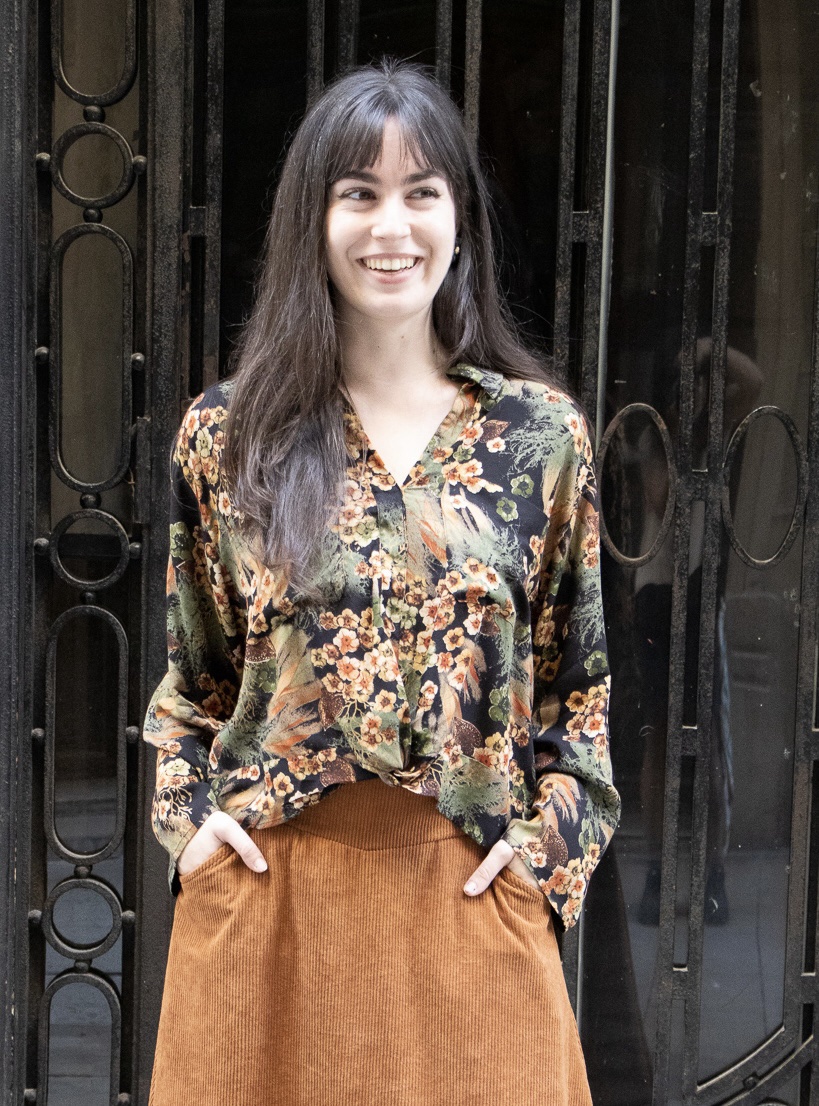 Mujer sonriendo frente a una puerta llevando una camisa con estampado marrón y naranja y una falda con bolsillos de pana color teja claro.