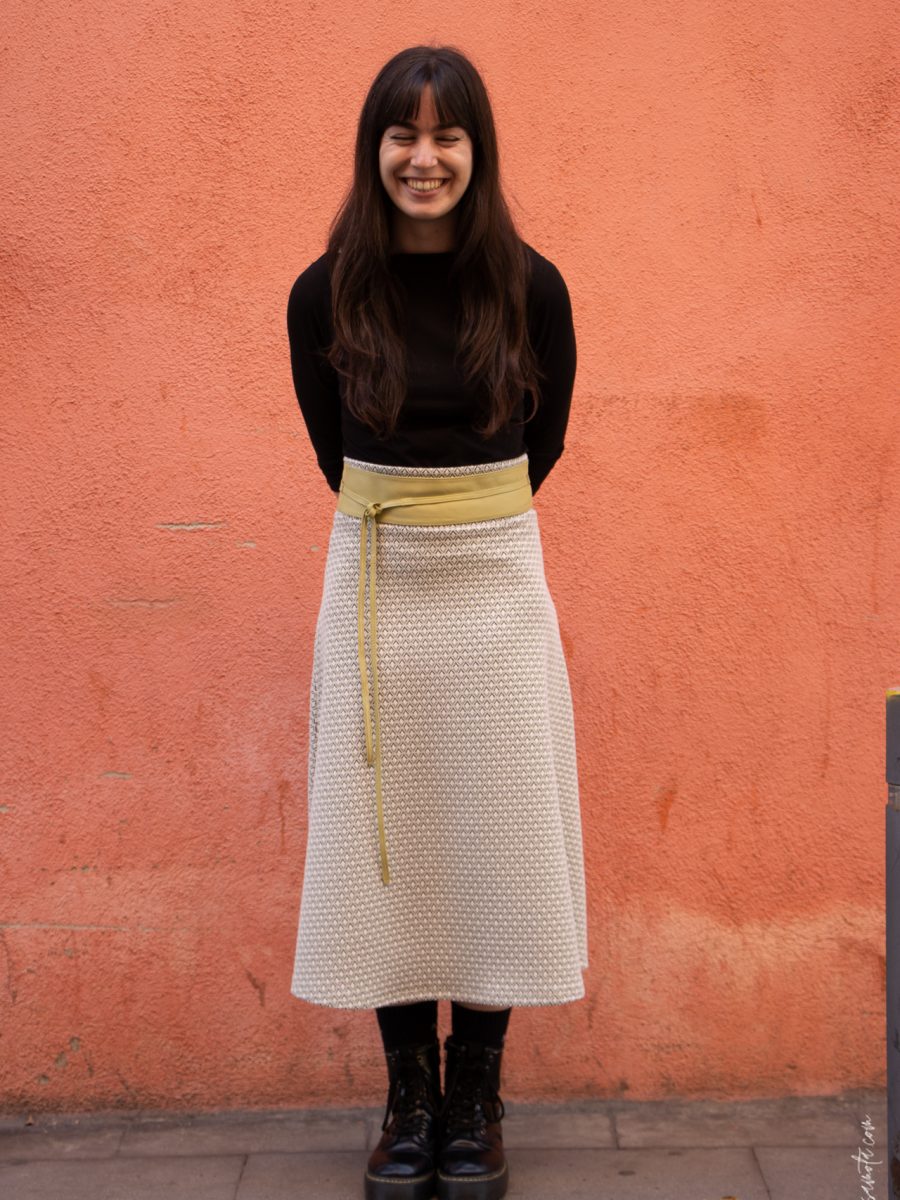 Mujer sonriendo en frente de una pared rojiza. Vestida en un top negro, una falda midi estampada, un cinturon amarillo y un par de botines negros.