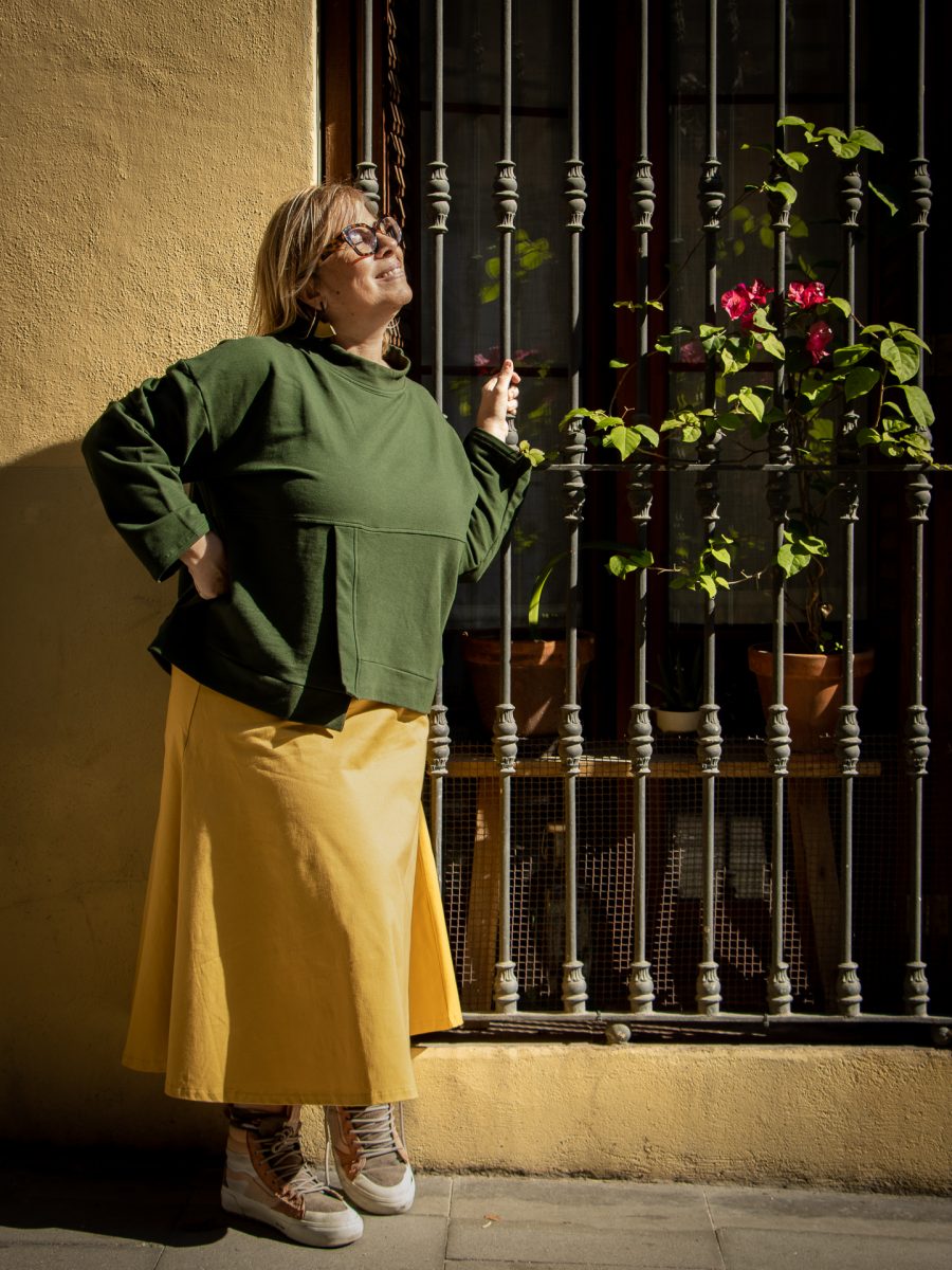 Mujer vestida con jersey verde botella, falda mostaza y zapatillas blancas con cordones rosados, de pie frente a una ventana con barrotes metálicos, disfrutando de la luz del sol. En el alféizar hay macetas con plantas verdes y flores rosadas, sobre una pared amarilla iluminada por sombras suaves.