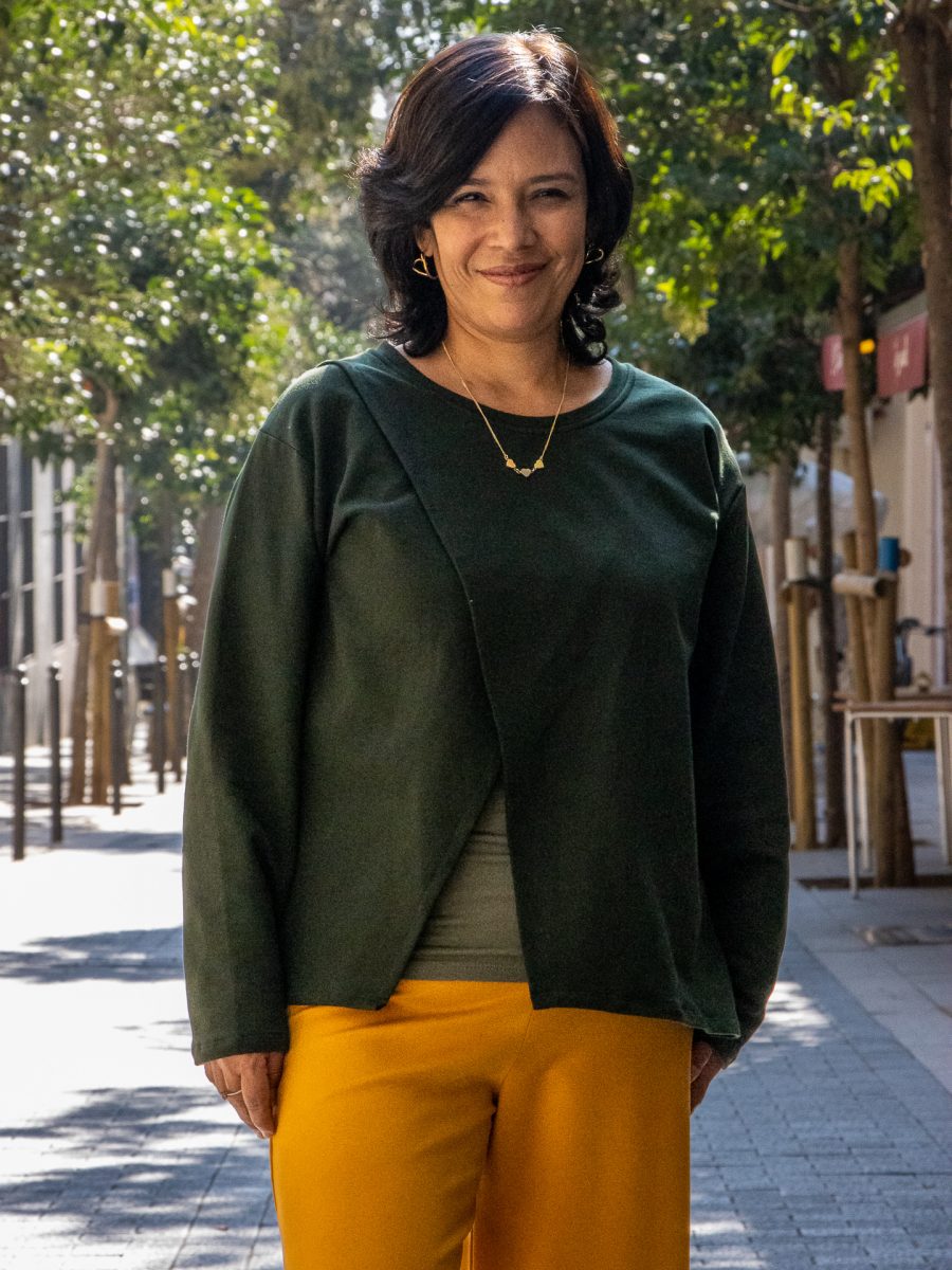 Mujer sonriente con Jersey verde botella de manga larga y diseño cruzado en el frente, combinada con pantalones amarillos, posando en un camino peatonal soleado rodeado de árboles y edificios con letreros al fondo.