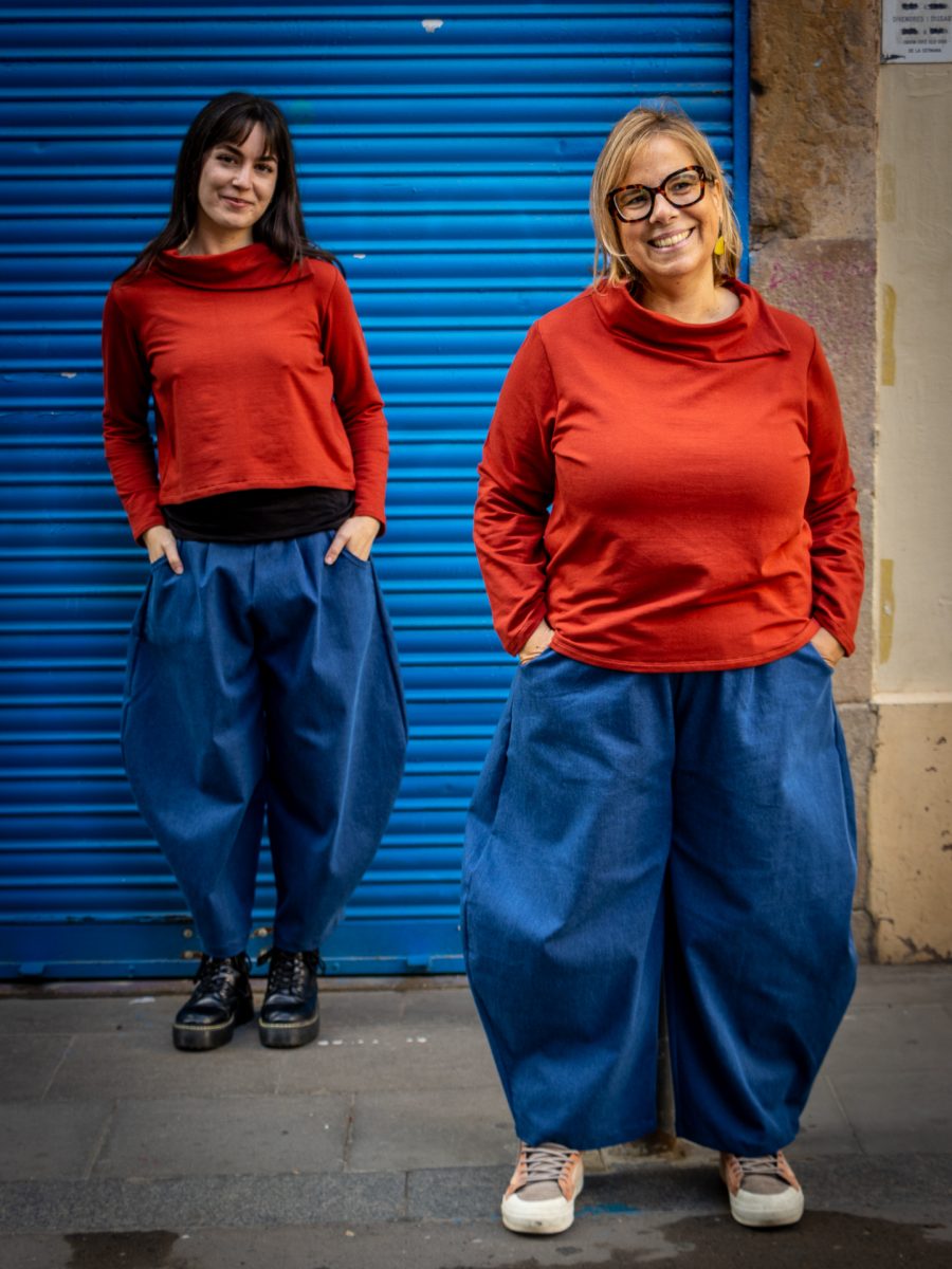 Dos mujeres con outfits coordinados —jerseys teja de manga larga y pantalones anchos de mezclilla azul— posan frente a una puerta metálica azul corrugada, creando un contraste vibrante con el fondo urbano desgastado.